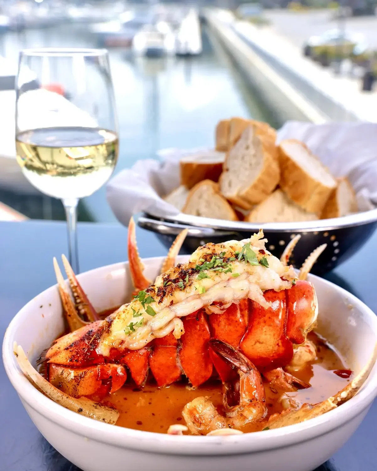 Killer Shrimp Lobster & Crab: lobster tail and snow crab claws in spicy sauce beside a glass of white wine and a basket of bread, marina view