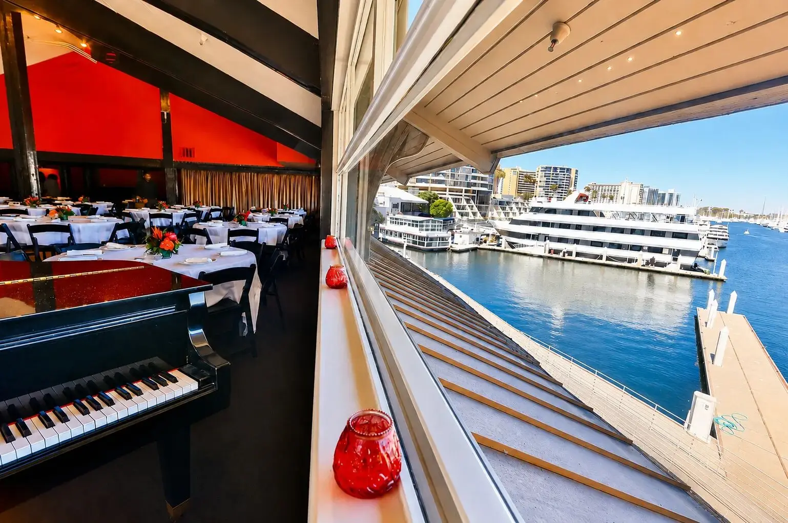 The Overkill Room private event venue set with white-linen banquet tables beside a grand piano, with windows looking out on yachts at the Marina del Rey docks
