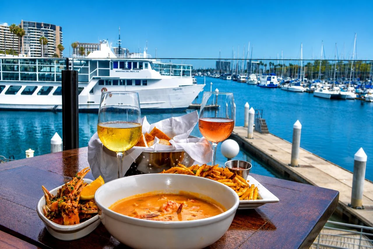 Bowl of Killer Shrimp in spiced orange broth with French bread, fries, and two glasses of wine on a waterfront table overlooking the Marina del Rey harbor and a docked yacht